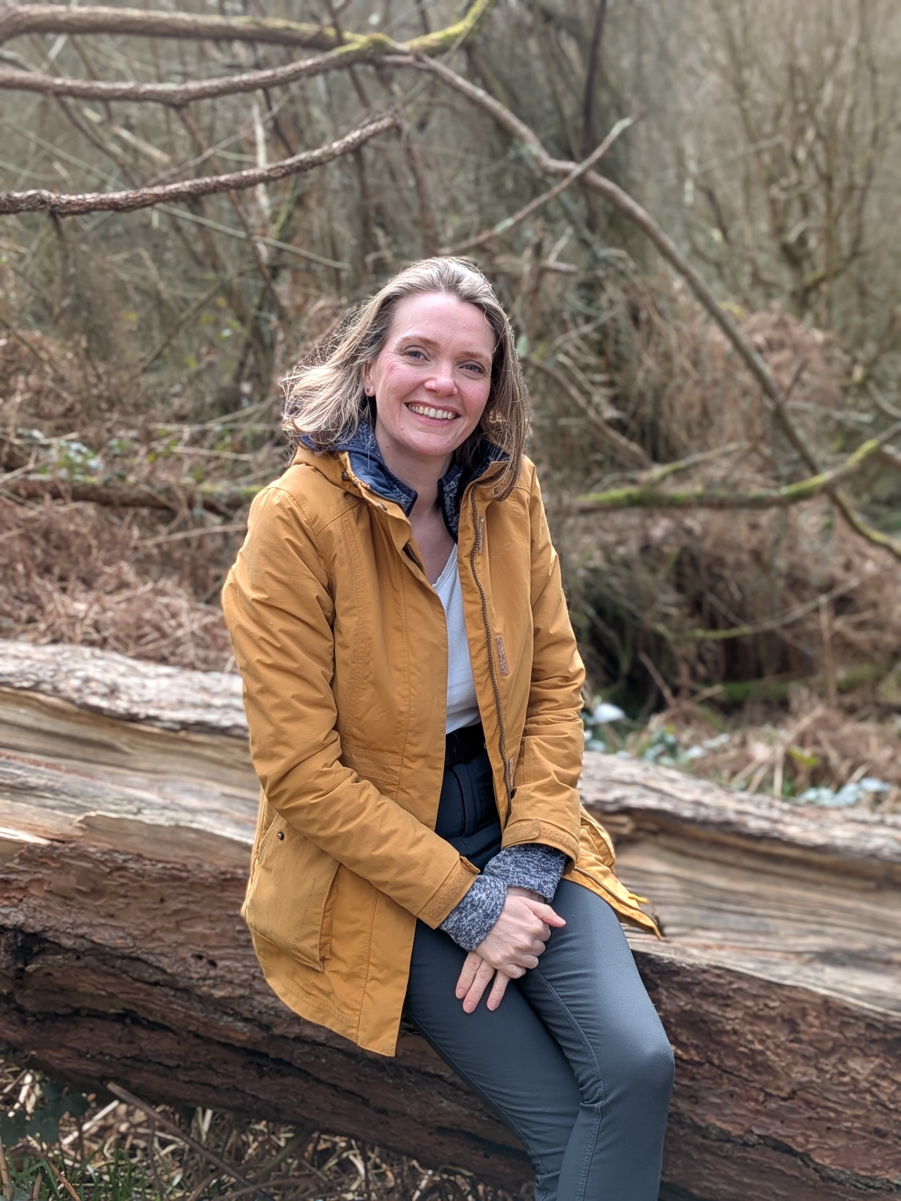 Emma Louise Morton, The Step in Time Celebrant. Sitting on a fallen log in the forest. Emma is smiling wearing a yellow coat.